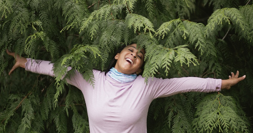 Woman standing in trees with her arms spread wide.