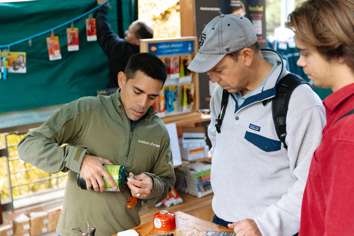 A founder demonstrates how to use a tool to open a can of propane to two customers in an open-air marketplace.